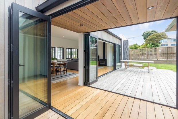 A bright contemporary indoor-outdoor living area with fully opening black-framed glazed doors connecting a timber-lined interior to a spacious timber deck. The interior includes a dining area and living room, while the deck overlooks a fenced suburban garden and includes a simple outdoor table.