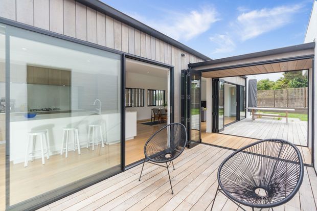 Contemporary single-level home opening onto a timber deck and sheltered courtyard. Large black-framed sliding glass doors connect the deck to a bright white kitchen with an island and stools, while a dining area and landscaped outdoor space are visible beyond. The facade features vertical light-toned cladding, dark metal framing, and a covered outdoor connection.