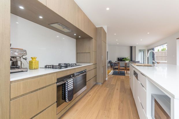 Bright contemporary open-plan kitchen with light timber cabinetry, white stone benchtops, integrated appliances, timber flooring, and a long central island. The kitchen opens toward a dining area and glazed outdoor connection at the rear.