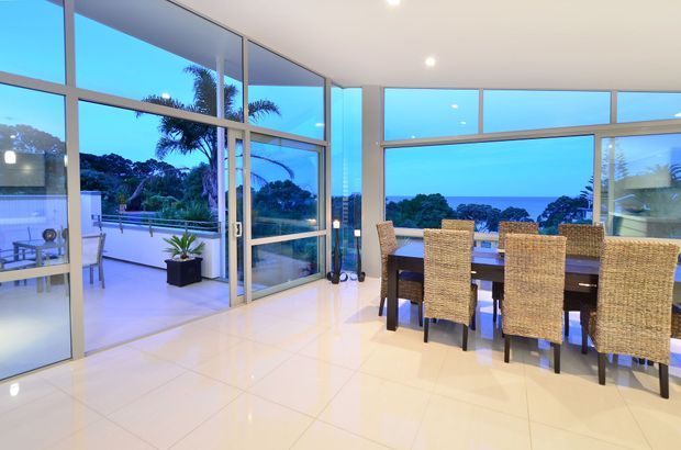 Bright contemporary dining area with a large dark timber dining table and woven high-back chairs, opening onto a terrace through extensive floor-to-ceiling glazing. The room has a glossy light tiled floor, white ceiling and walls, slim aluminium-framed glass doors and windows, and expansive views of lush vegetation and blue water beyond.