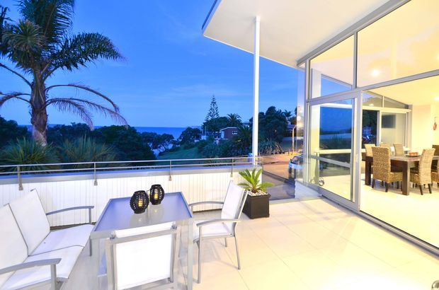 Bright contemporary outdoor terrace overlooking a coastal landscape, furnished with a white outdoor dining table and chairs. The terrace has pale tiled flooring, white slatted privacy panels, stainless steel or aluminium balustrading, and a broad glazed opening into a warmly lit dining room. Palm trees and ocean views are visible beyond.