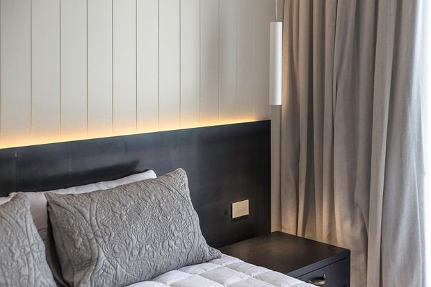 Close-up view of a contemporary bedroom corner featuring a bed with textured neutral bedding, a dark built-in headboard extending into a bedside table, subtle integrated LED backlighting, vertically panelled light walls, and full-height taupe curtains beside a window. The composition is calm and refined with a restrained neutral palette.