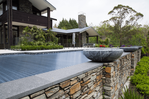 Exterior view of a modern multi-level residence beside a large infinity-edge swimming pool. The house features dark horizontal cladding, extensive glazing, a pitched roof, and a prominent rough-stone chimney. A stacked natural-stone retaining wall with integrated sculptural planters borders the pool, surrounded by landscaped vegetation.