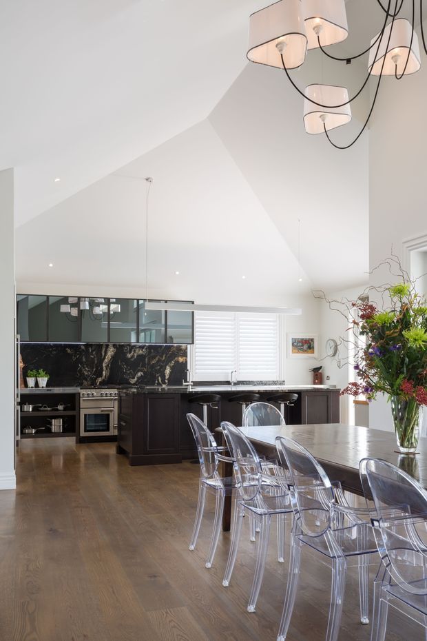 A bright, open-plan kitchen and dining area with a dramatic vaulted white ceiling. The kitchen features dark timber cabinetry, a black marble-look splashback and benchtop, stainless steel appliances, and a long island with bar seating. A large dining table with transparent acrylic chairs sits in the foreground beneath a sculptural multi-pendant light. Timber flooring, extensive glazing, and white plantation shutters complete the space.