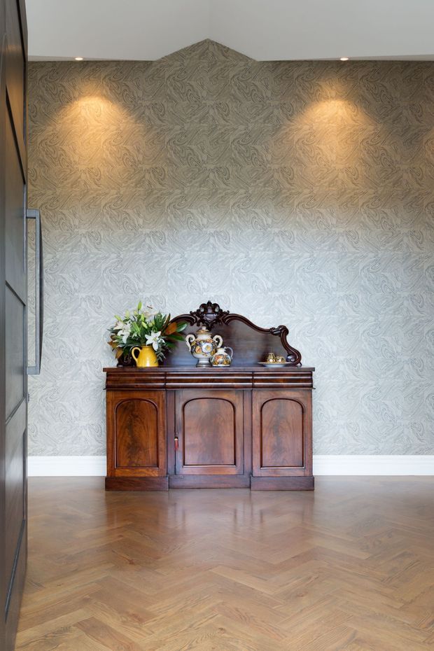 A refined interior vignette featuring an ornate dark timber antique sideboard with decorative mirror-back detail and porcelain objects, positioned against a textured grey patterned feature wall. The space has warm recessed lighting, white skirting, and a herringbone timber floor, with a partial dark-framed glazed opening visible at left.