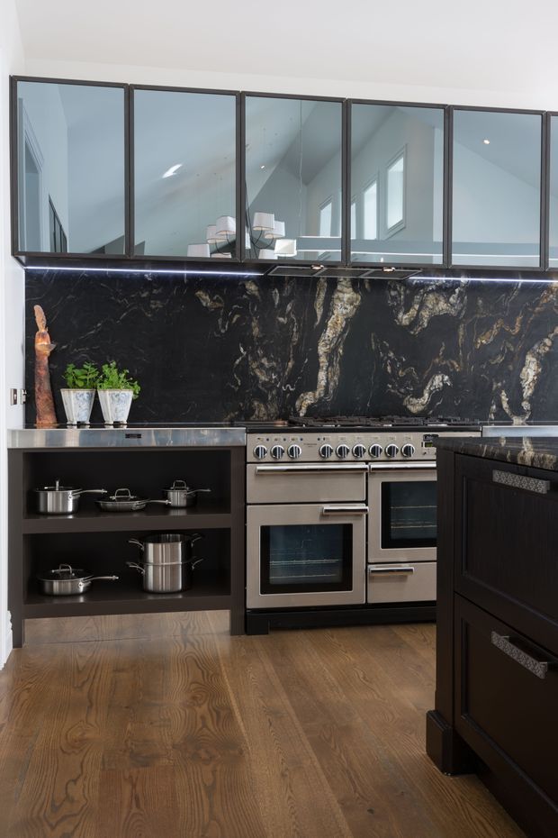 A sophisticated kitchen featuring dark cabinetry, a large stainless-steel range cooker, dramatic black stone backsplash and benchtops with prominent light veining, and warm timber flooring. Upper mirrored cabinet doors reflect the bright contemporary interior and pendant lights. Open shelving displays stainless-steel cookware, while integrated lighting highlights the backsplash.