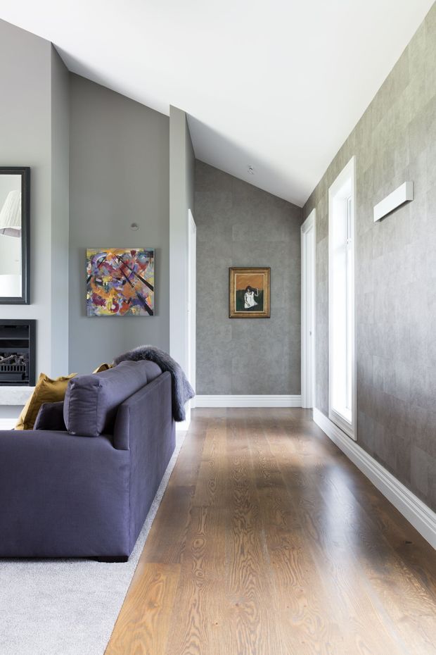 A bright, narrow interior passage connecting to a living room. The space features warm timber flooring, grey textured walls, white trim and doors, tall glazing along one side, and a partially visible deep purple sofa with artwork and a mirror. The sloping ceiling and angled wall junctions create a contemporary architectural character.
