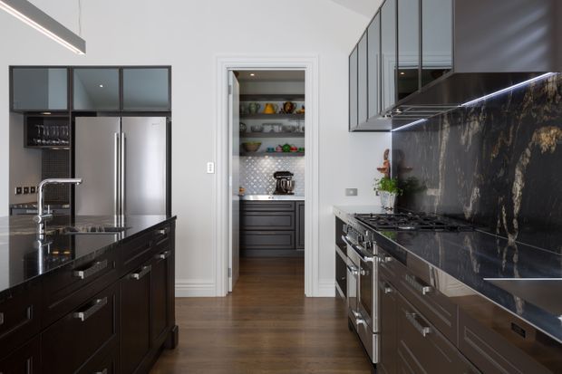 A sophisticated galley-style kitchen with dark timber cabinetry, a large central island, stainless-steel appliances, and a dramatic dark stone splashback and benchtop. Glass-front upper cabinetry and a walk-in pantry with open shelving are visible at the rear. The room has a restrained, high-contrast palette of charcoal, white, stainless steel, and warm timber flooring.