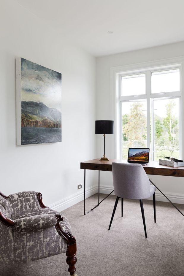 Bright, refined home office with white walls, a large divided window, a slim dark timber desk positioned beneath the window, a laptop, black table lamp, upholstered modern chair, and a traditional carved wooden armchair. A large landscape artwork provides a muted blue-grey accent. The room has carpeted flooring and classic wall and window trim.