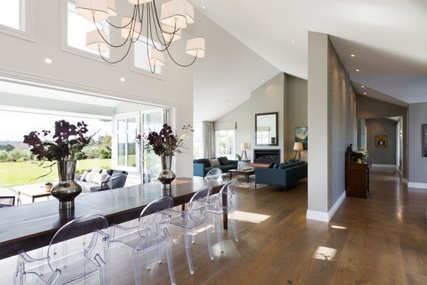 Bright open-plan interior showing a dining area in the foreground with a long dark wood table and transparent acrylic chairs, opening into a living room and hallway. The space features warm timber flooring, white walls, extensive glazing, high angled ceilings, recessed lighting, and a large contemporary chandelier. A landscaped garden is visible through the open doors and windows.
