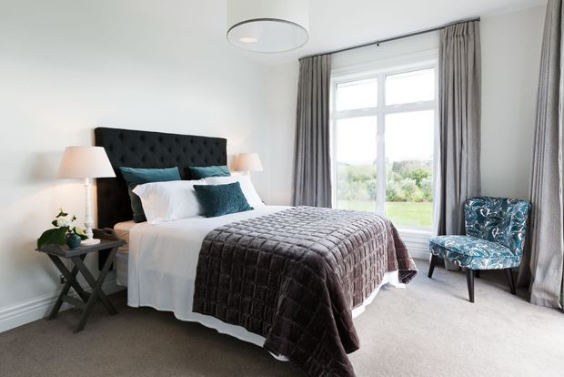 A bright, refined bedroom with a black tufted upholstered headboard, layered white bedding, and a dark quilted bedspread. Floor-to-ceiling grey curtains frame a large white-trimmed window, while a patterned blue accent chair, bedside tables, lamps, and a circular ceiling light complete the room.