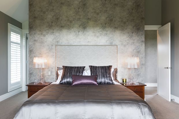 A refined bedroom viewed straight on, centred on a large upholstered bed with dark satin bedding, patterned cushions, and matching bedside tables with lamps. The room features a textured grey feature wall, light upholstered headboard, grey painted walls, carpeted floor, white plantation shutters, and an open white door.