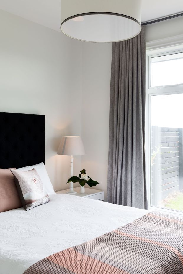 Bright, softly styled bedroom with a black upholstered headboard, white textured bedding, blush and neutral cushions, and a patterned blush-grey throw. A white bedside table holds a small lamp and plant, while full-height grey curtains frame a large white-framed window. A large cylindrical pendant light hangs overhead, creating a calm contemporary interior.