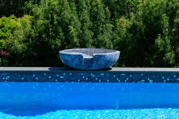 Close-up exterior view of a vivid blue swimming pool with a mosaic-tiled coping and a large sculptural bowl water feature positioned along the far edge. Dense green landscaping forms the background.