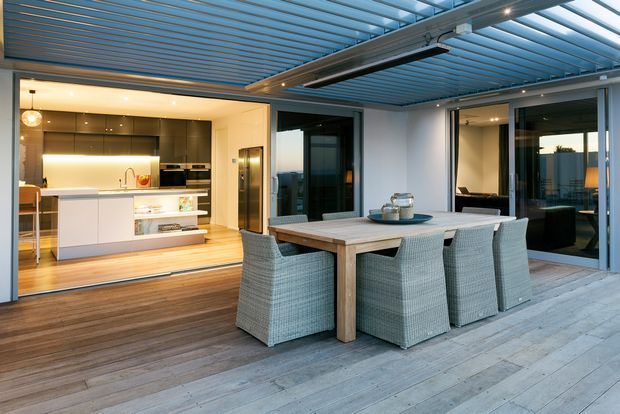A contemporary covered outdoor dining terrace with a large timber dining table and four woven dining chairs, adjoining an illuminated modern kitchen through wide glazed openings. The terrace has weathered timber decking, a translucent roof with exposed metal framing, integrated linear lighting, and dark-framed sliding glass doors.