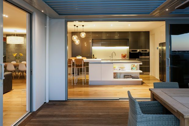 A contemporary indoor-outdoor entertaining area opens directly into a bright kitchen and dining space. The kitchen features a central white island with integrated shelving and seating, dark cabinetry, timber flooring, pendant lighting, and large sliding glass doors. In the foreground, a timber deck has an outdoor dining table and woven chairs beneath a slatted overhead cover. A dining room is visible through the opening at left.