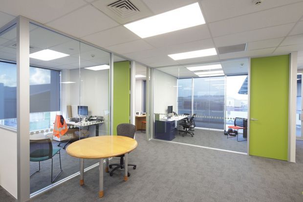 Bright modern commercial office interior with an open circulation area, glass-partitioned workspaces, lime-green doors and wall panels, grey carpet, white suspended ceiling with recessed fluorescent lighting, desks and office chairs, and large windows overlooking an urban setting.