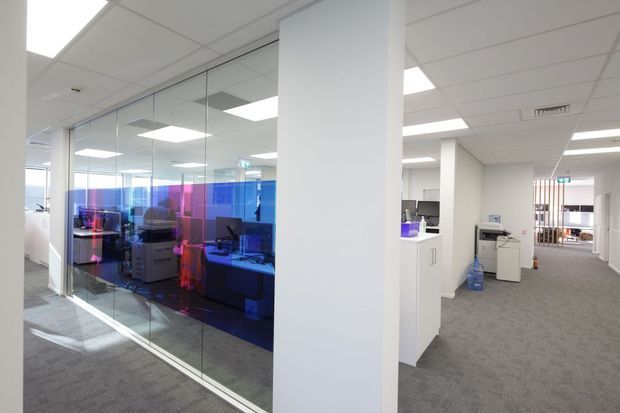 Bright contemporary commercial office interior with an open-plan workstation area enclosed by full-height glass partitions. The office features white structural columns and walls, a suspended acoustic tile ceiling with recessed fluorescent lighting, grey carpet, white cabinetry and workstations, and vivid blue and magenta translucent privacy film on the glass. A corridor with printers and office equipment extends into the background.