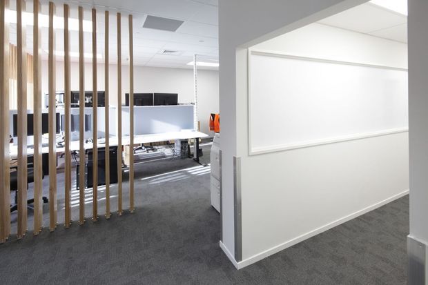 Bright contemporary commercial office interior viewed from a carpeted corridor. An open-plan workstation area is partially screened by vertical timber battens, with white desks, computer monitors, office chairs, and storage visible beyond. The corridor features white walls with a large recessed rectangular wall panel and grey carpet flooring.