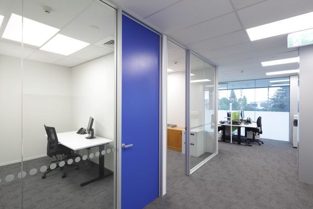 A bright contemporary office interior with a carpeted open-plan workspace, enclosed meeting room, glass partition walls, and a vivid blue solid door. Desks, office chairs, computer equipment, and timber-toned storage are visible beneath a suspended acoustic tile ceiling with recessed fluorescent lighting.