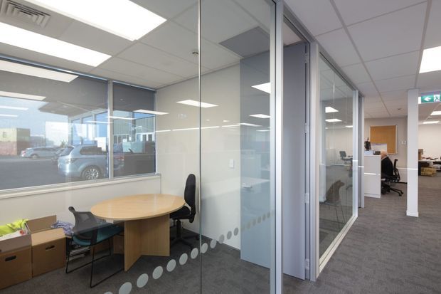 Bright contemporary commercial office interior showing a carpeted corridor lined with glazed meeting rooms and private offices. The foreground meeting room has a round timber table, office chairs, fixed glass partitions with frosted dot decals, and a suspended grid ceiling with recessed fluorescent lighting. Workstations and office furniture are visible through the glazing.