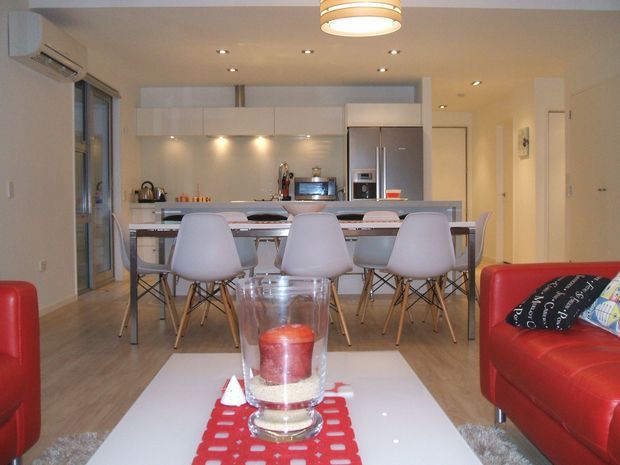 Open-plan contemporary interior combining a kitchen, dining area and living room. The kitchen has white flat-panel cabinetry, integrated under-cabinet lighting, stainless-steel appliances and a long island or breakfast counter. A dining table with white molded chairs sits between the kitchen and the living area, while red leather sofas and a white coffee table occupy the foreground. Recessed ceiling lights, timber-look flooring and large glazed doors create a bright, functional space.