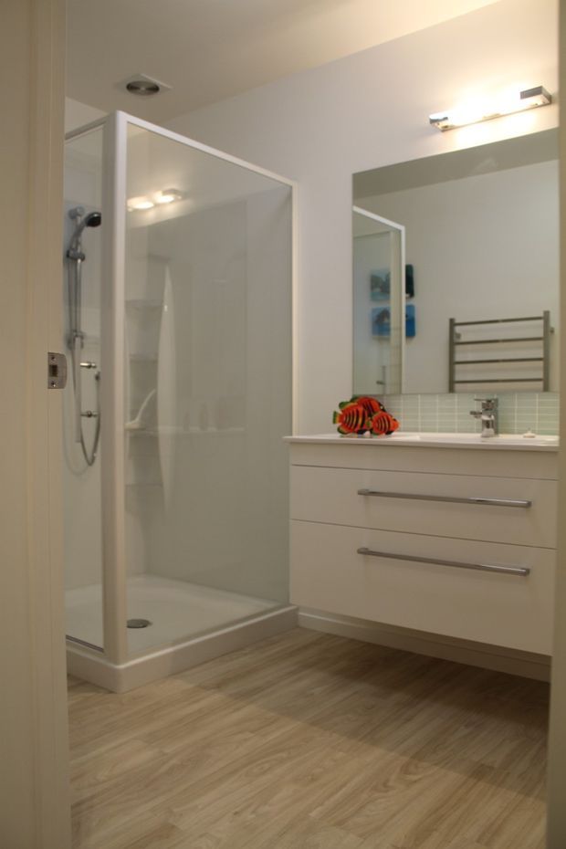 A clean, contemporary bathroom viewed from the doorway, featuring a large framed glass shower enclosure with handheld shower fixture, floating white vanity with long metal handles, wall-mounted basin tap, pale green small-tile splashback, large mirror, heated towel rail, and warm wood-look flooring.