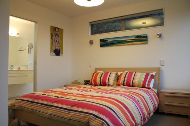 A compact, warmly lit bedroom with a double bed dressed in bright multicoloured striped bedding. The room has pale walls, a narrow high window, timber bedside furniture, ceiling lighting, and colourful wall artwork. An open doorway reveals a bathroom with a vanity and mirror.