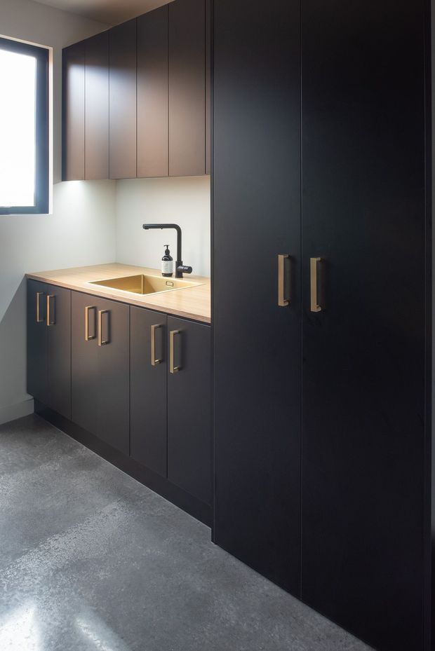 Compact contemporary utility or kitchenette area with floor-to-ceiling matte dark cabinetry, integrated storage, a pale timber-look benchtop, undermount sink, and black gooseneck tap. A small window provides natural light, while the polished grey concrete floor adds an industrial note. Brass-toned handles create a subtle accent.