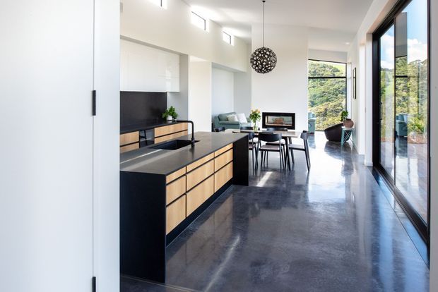 Bright open-plan contemporary interior with a black kitchen island featuring light timber drawer fronts, polished concrete flooring, a dining area, living seating, and expansive floor-to-ceiling glazing opening toward a leafy landscape. A sculptural spherical pendant provides a strong decorative focal point.