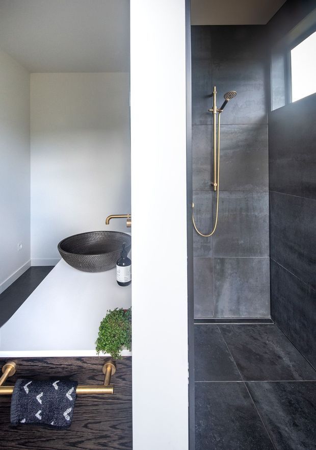 Contemporary bathroom with a walk-in shower enclosed by a partial white wall. The shower features large dark grey stone-look tiles, matching dark floor tiles, a high horizontal window, and brushed brass shower hardware. In the adjacent vanity area, a white countertop supports a black textured vessel basin, brass tapware, soap dispenser and greenery, with a dark timber vanity front and brass towel rail.