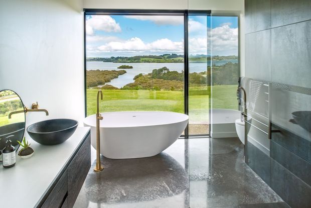 Contemporary bathroom with a freestanding white bathtub positioned in front of expansive floor-to-ceiling glazing overlooking a landscaped lawn, water, and greenery. The room includes a dark vessel basin on a timber vanity, a brass floor-mounted bath filler, large grey tiled walls, and a polished grey floor. The design is bright, refined, and strongly connected to the surrounding landscape.