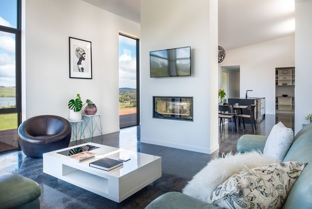 Bright contemporary living room with floor-to-ceiling glazing, a wall-mounted television above a built-in fireplace, a white geometric coffee table, curved leather lounge chair, and a pale sectional sofa. A dining area and kitchen joinery are visible beyond the open transition. The room has polished dark flooring, white walls, and expansive views through the windows.
