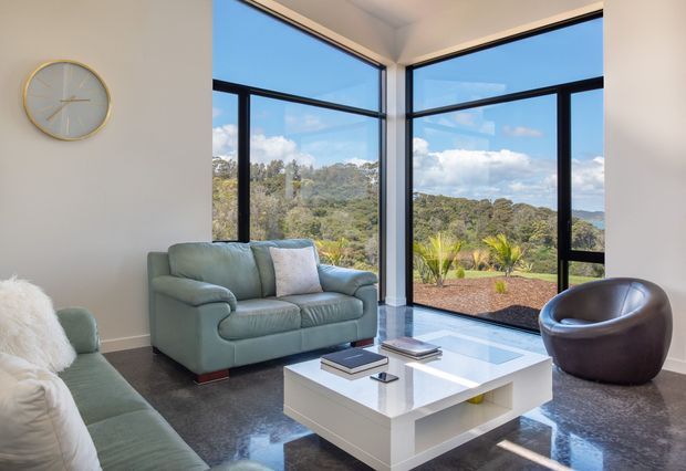 Bright contemporary living room with a pale green sofa and armchair, white glossy coffee table, dark polished flooring, and expansive black-framed corner glazing overlooking a wooded landscape. A simple round wall clock and minimal furnishings contribute to a clean, refined interior.