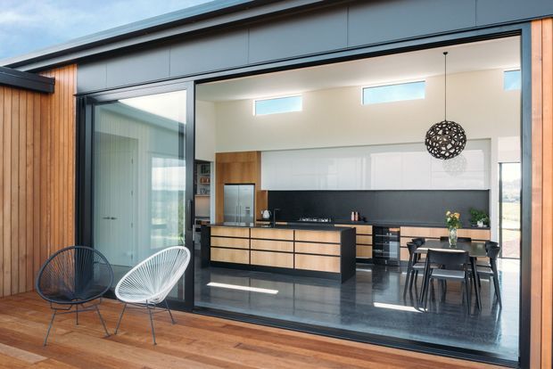 Contemporary open-plan kitchen and dining area viewed from a timber deck through large sliding glass doors. The interior features dark polished flooring, a long timber-and-black kitchen with island, white cabinetry, clerestory windows, a dining table, and sculptural pendant lighting. Vertical timber wall cladding and outdoor lounge chairs frame the exterior view.