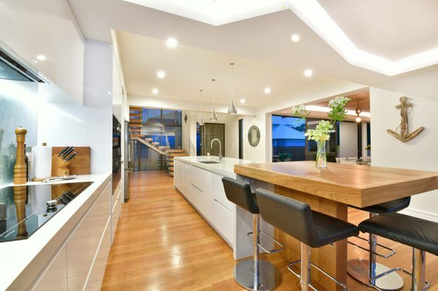 Bright open-plan kitchen and dining area with a long white island, integrated appliances, timber flooring, and a substantial timber dining bench or table with black bar stools. Recessed and cove lighting, pendant lights, reflective splashback surfaces, and nautical wall decor create a polished contemporary coastal feel. A staircase and adjoining living area are visible beyond the kitchen.