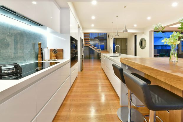 Bright contemporary open-plan kitchen with extensive white cabinetry, a long island with an integrated sink, timber benchtop and bar seating, polished timber flooring, pendant lights, and a dark stone splashback. A dining or living area and stairway are visible beyond the kitchen.