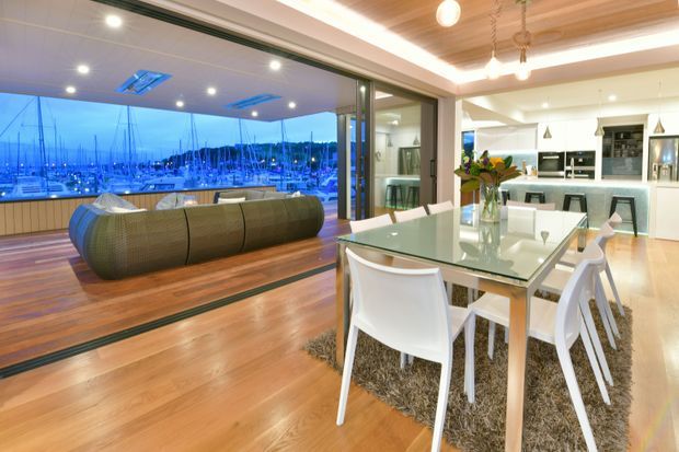 Open-plan contemporary dining and living area with warm timber flooring and a timber-lined recessed ceiling. A glass dining table with white chairs sits beside a modern kitchen with white cabinetry and dark benchtops. Full-height sliding glass doors open to a marina view, with a curved sofa positioned in the adjacent living zone.