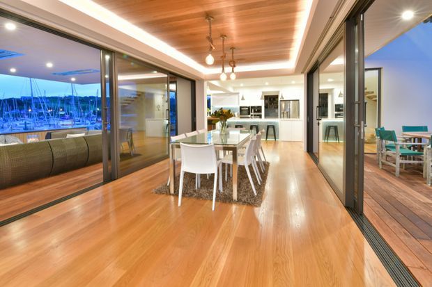 Bright open-plan dining and kitchen interior with warm timber flooring and a timber-lined recessed ceiling. Large dark-framed sliding glass doors open to a balcony or terrace overlooking a marina, with additional outdoor dining visible on the right. A white dining table and chairs sit on a textured rug in the foreground, while a contemporary white kitchen is visible beyond.
