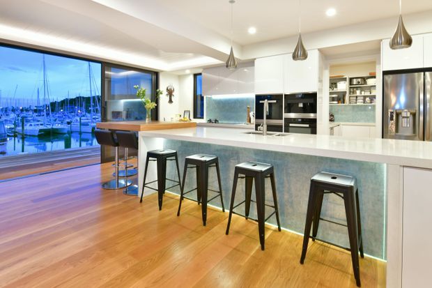 Bright contemporary open-plan kitchen with a long white island and integrated breakfast bar, five dark metal stools, glossy white cabinetry, stainless-steel appliances, and a softly illuminated blue-grey stone-look island face. Warm timber flooring and pendant lights add contrast, while expansive glazing provides a marina view.