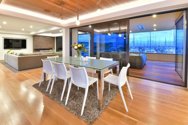 Open-plan contemporary dining and living area with warm timber flooring and a timber-lined ceiling. A glass dining table with white chairs sits on a textured rug, while the adjoining living room includes a sectional sofa and fireplace. Full-height glazed doors open to a timber deck with a marina and boats visible beyond.