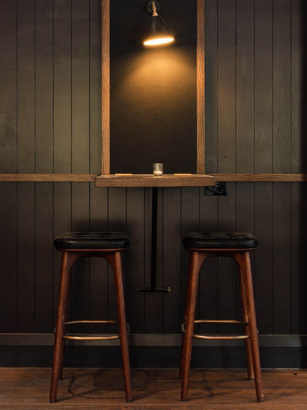 A dark, intimate bar-style interior with two wooden stools positioned beneath a narrow timber counter. Vertical dark wall panelling frames a central recessed niche with a warm pendant light, small shelf, and tap-like fixture. The space has a moody, rustic-industrial character with timber flooring and black metal details.