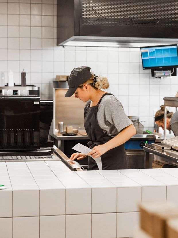 A commercial food-service kitchen with white tiled walls and counters, stainless-steel equipment, dark built-in cabinetry or ventilation above, and a worker operating coffee or kitchen equipment while holding a printed order. Another staff member and food-service equipment are visible in the background.