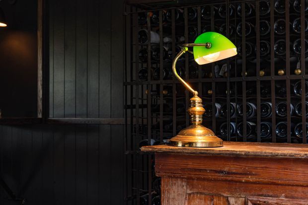 Moody close-up of a vintage green-shaded desk lamp with a brass-colored stem and base, positioned on a richly patinated timber counter. Behind it is a dark metal wine-bottle storage rack filled with bottles, while dark vertical wall panelling reinforces the intimate, traditional wine-cellar atmosphere.