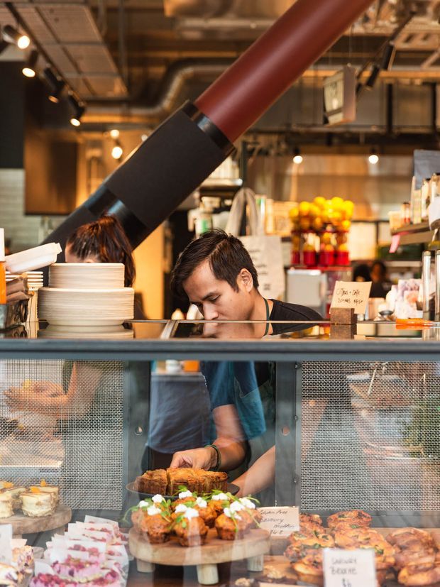 A warmly lit industrial-style bakery or café counter, with staff preparing and displaying pastries behind glass. The space features exposed services, metal framing, hanging track lights, a large red pipe, and open commercial kitchen shelving.
