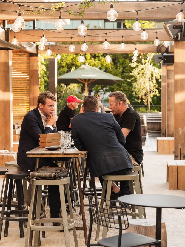 An open-air hospitality venue with timber pergola beams, warm wood screening and numerous suspended globe string lights. Diners sit at tall communal tables and stools, with additional café seating, an outdoor umbrella and lush greenery visible beyond.