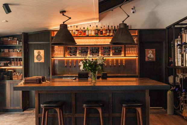 A warmly lit, dark-toned cocktail bar interior with a large timber-topped counter and three stools in the foreground. Behind it are black framed wall panels, illuminated liquor shelves, a back bar, pendant lights, chalkboard menus, and open shelving displaying bottles and glassware. The space has a refined, atmospheric hospitality design with strong amber accent lighting.