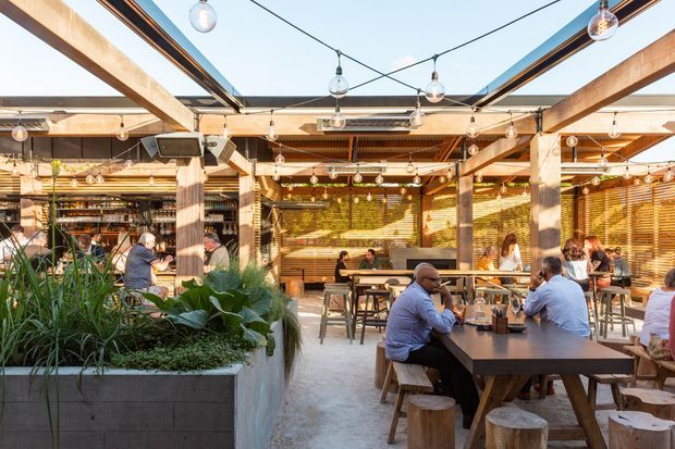 A bright, open-air commercial dining area with timber posts and beams, exposed steel framing, overhead string lights, timber Venetian-style screens, concrete planters, and communal timber tables with stools. Diners occupy the space, which has a relaxed contemporary hospitality atmosphere and views of the blue sky through the open roof structure.