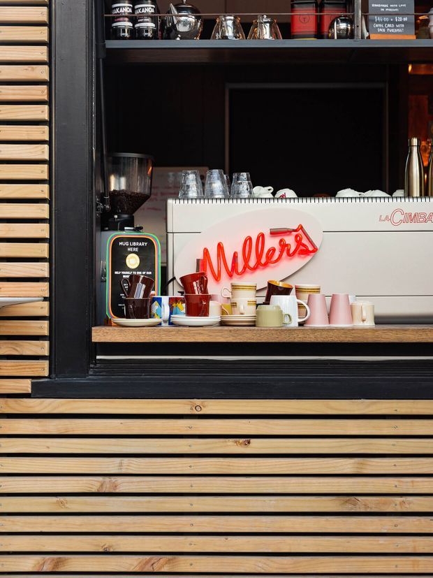 Close-up of a coffee service window set into a dark-framed timber-clad facade. A white La Cimbali espresso machine, grinder, colourful cups, jars, and a red neon “Millers” sign sit on a timber counter, with open shelving above displaying coffee equipment and supplies.