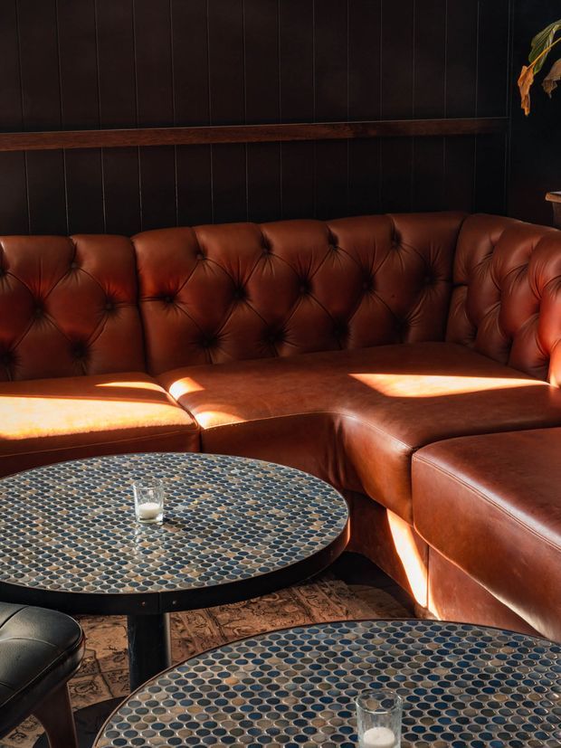 A moody lounge or hospitality seating area with a large cognac-brown tufted leather sectional wrapping around the corner. Two round tables have dark metal edges and patterned mosaic-like blue-grey tops, with a small glass visible on each. The room features dark vertical wall panelling, a horizontal timber trim, warm sunlight, and a partially visible plant, creating an intimate vintage-inspired atmosphere.